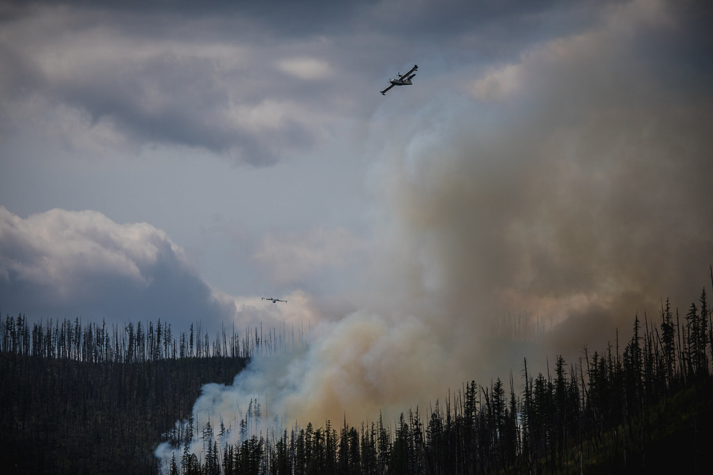 Fire suppression aircraft drop water near a wildfire with smoke over a mountain landscape.
