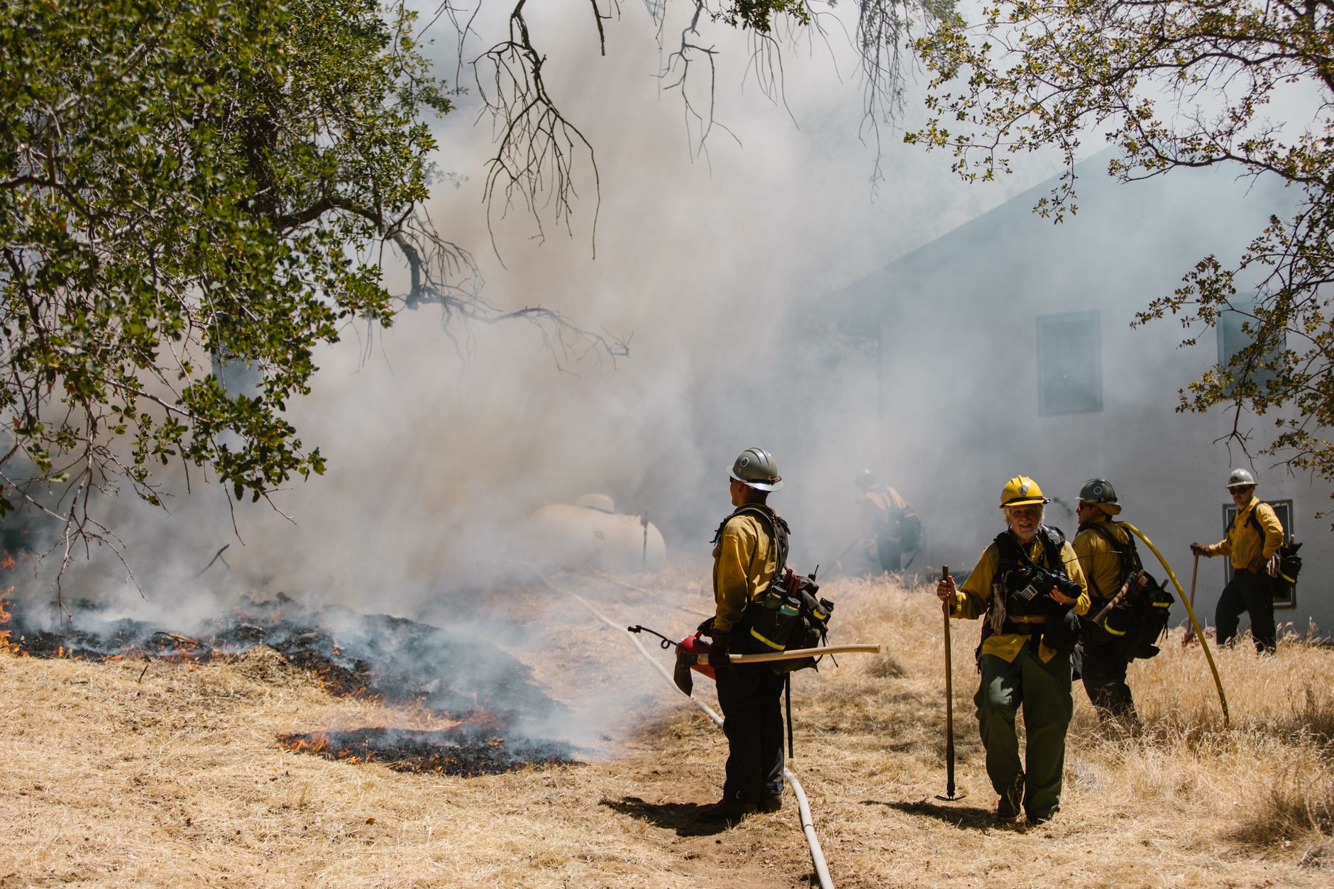 Firefighters in protective gear working near a smoky grass fire.