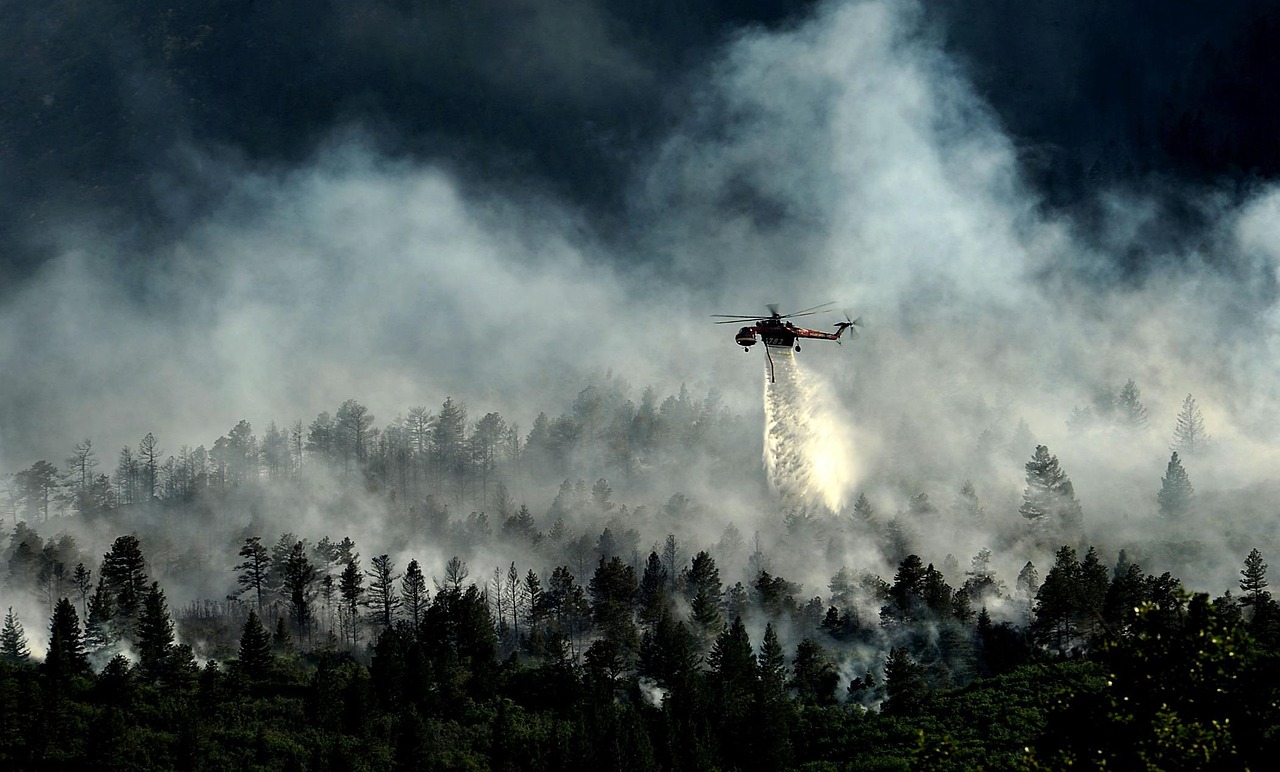 A helicopter flies above a forested landscape with wildfire smoke visible in the sky.