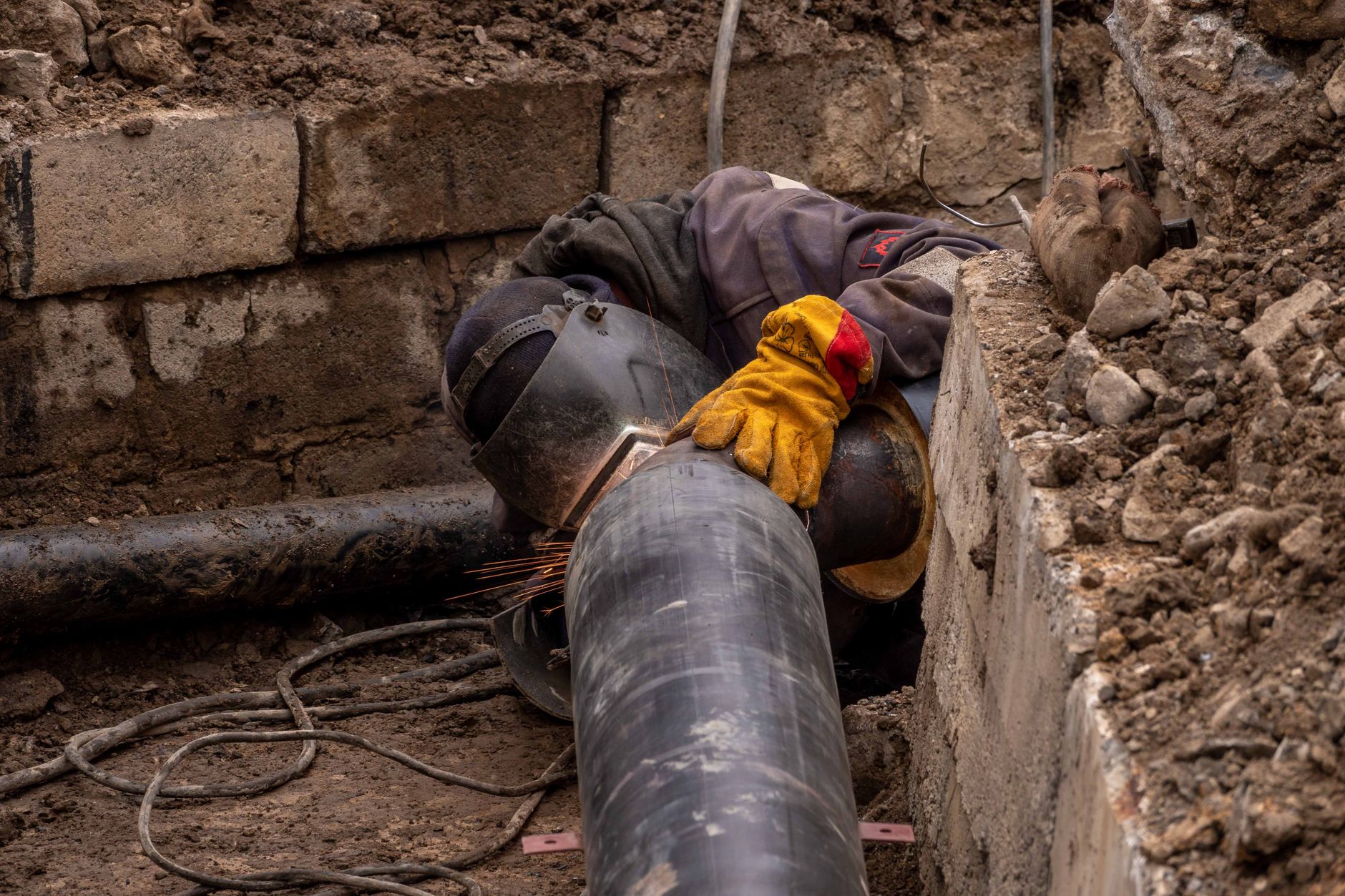 A worker welds a large pipe in an urban trench.